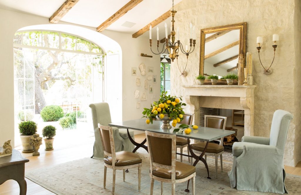 High ceiling white dining area with exposed wood beams, white fireplce and mantle, metal topped dining table with lemons, french doors in archway looking out to garden with large oak tree.